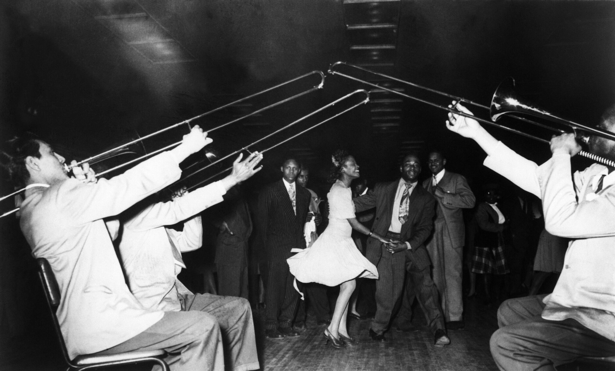 An African-American couple dances in the background to the Swing band playing their trombones in the foreground